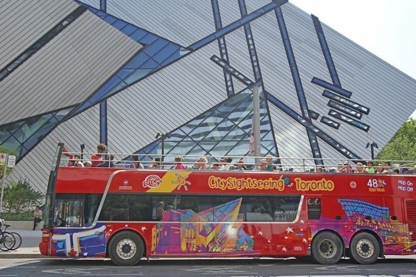Een kleurrijke dubbeldekker met 'City Sightseeing Toronto' erop, geparkeerd voor een modern gebouw met een glazen gevel. 1