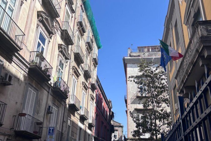 A narrow street with multi-story buildings, a clear blue sky, and a flag with a tricolor design. 2