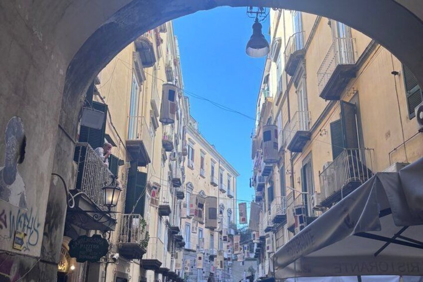 A narrow street with buildings on either side, a clear blue sky, and hanging laundry. 3