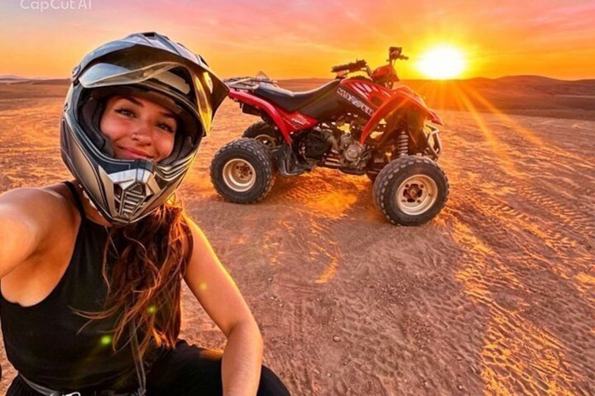 A person in a helmet sitting next to a red ATV on a desert road with the sun setting in the background. 1