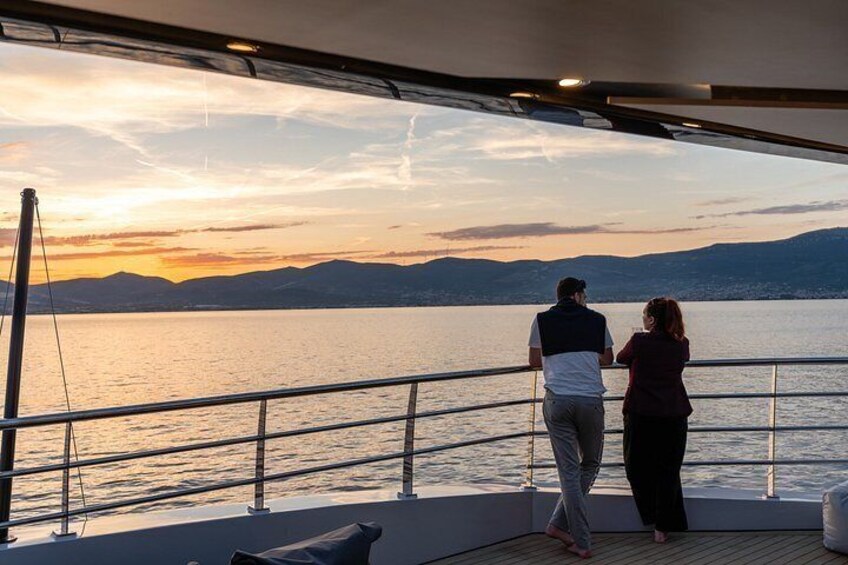 Un couple se trouve sur le pont d’un bateau, surplombant une étendue d’eau calme, avec un coucher de soleil en arrière-plan. 2