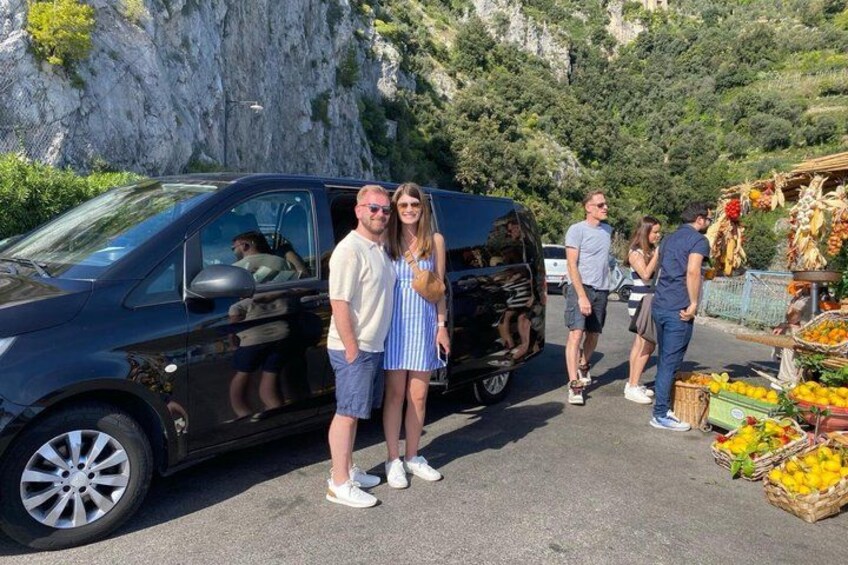 Un gruppo di persone in piedi vicino a un furgone nero e a un mercato di frutta. 2