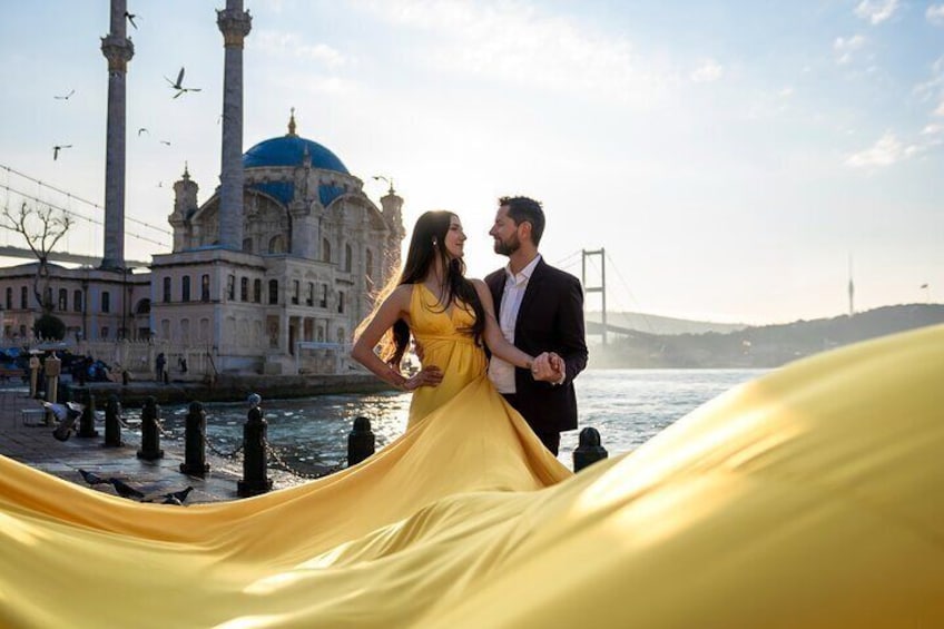 A couple in a yellow dress and black suit by a waterfront with a mosque and a bridge in the background. 1