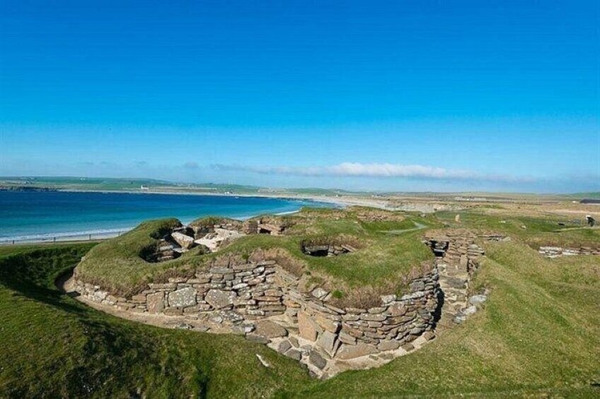 A coastal archaeological site with stone structures and grassy mounds. 1