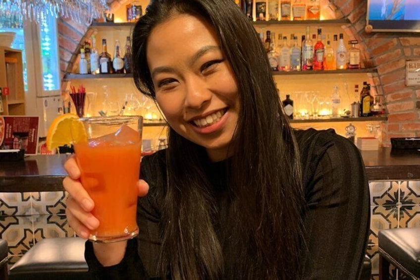 A person holding a glass with a red beverage, a bar with shelves of bottles, and a patterned wall in the background. 1