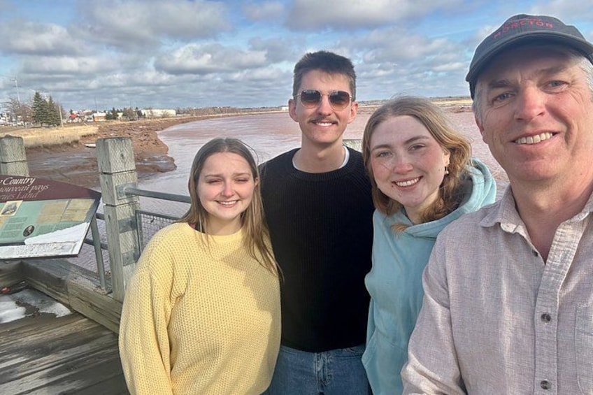 A group of people standing on a wooden deck with a river and a cloudy sky in the background. 1