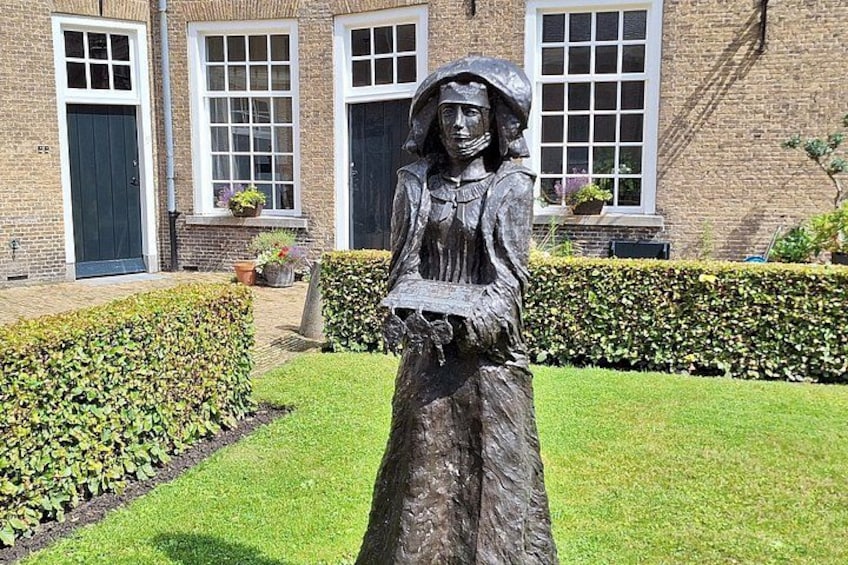 A statue of a woman in a dress and bonnet in a grassy area with a brick building in the background. 2