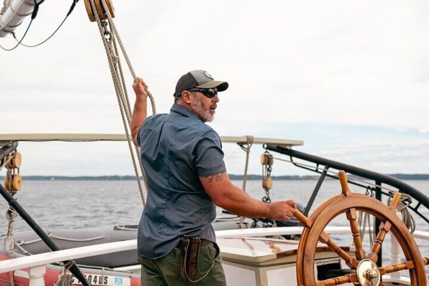A man steering a boat with a wooden wheel, ropes, and a calm sea in the background. 4