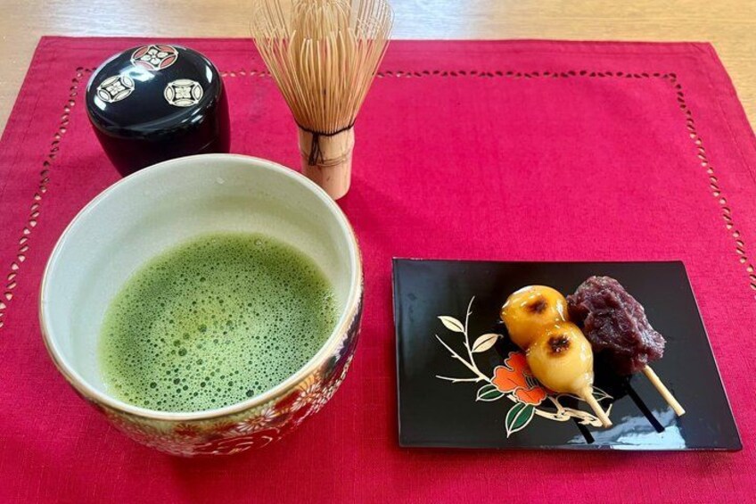 A cup of green tea, a black teapot, a whisk, and a plate with food on a red tablecloth. 2