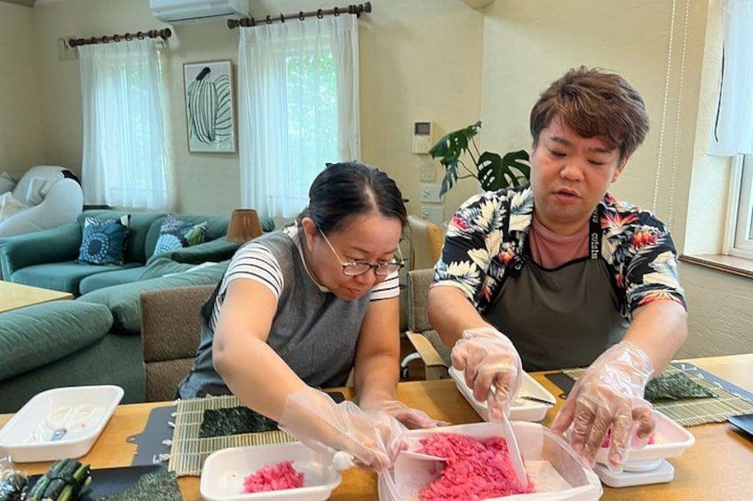 Two people wearing gloves, working with pink materials on a table in a room with a couch and curtains. 5