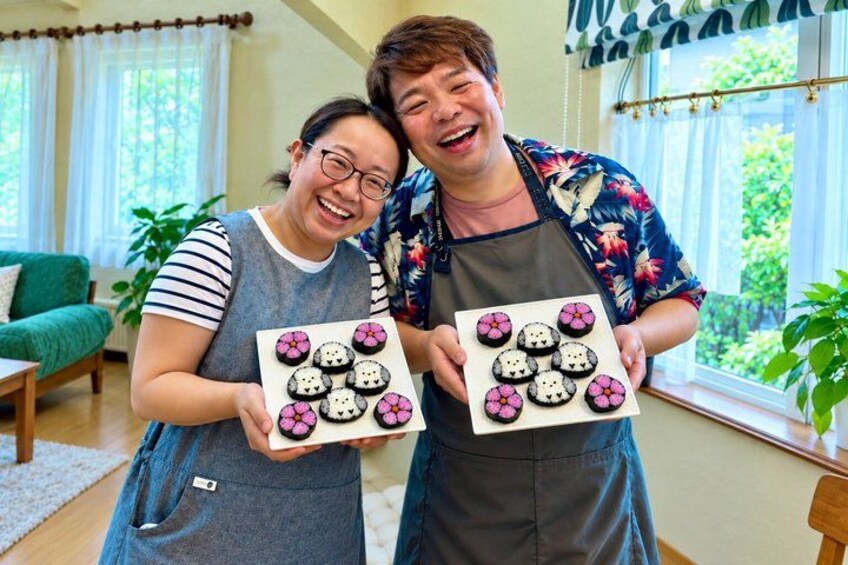 Two people holding plates of decorated food items in a room with a green sofa, a window with curtains, and a plant. 1