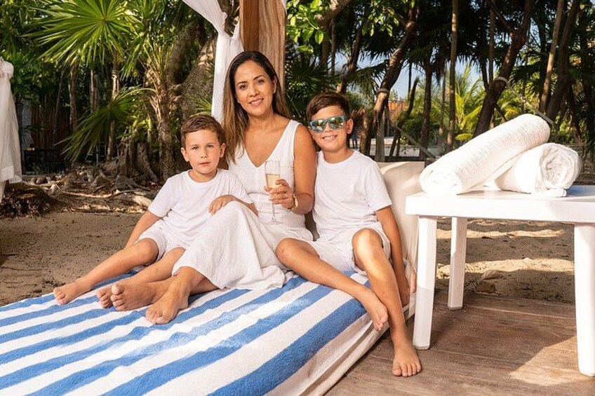 En familie på tre på en strand med en palme og et hvidt bord med håndklæder. 2