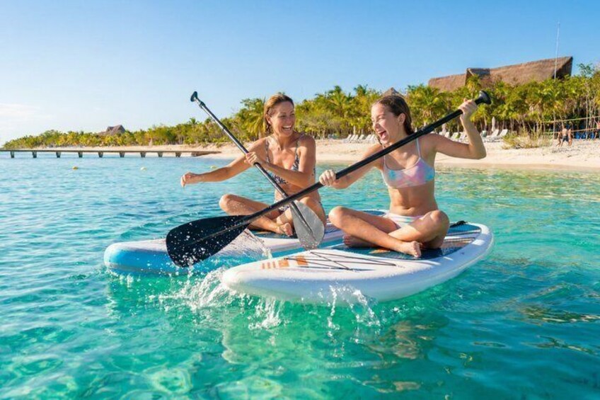 Due persone che fanno paddleboard su un mare turchese calmo, con una spiaggia e palme sullo sfondo. 1