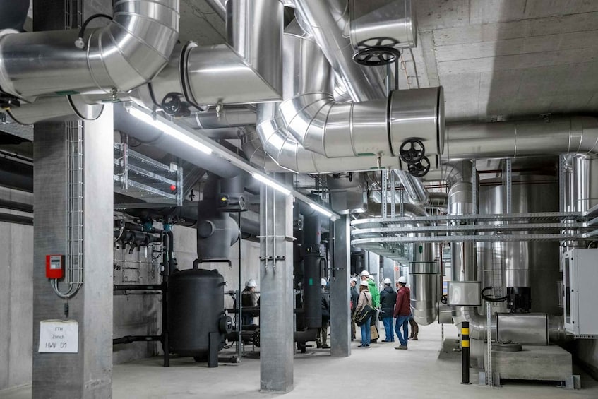 An industrial facility with large pipes, tanks, and machinery, and people observing the equipment. 1