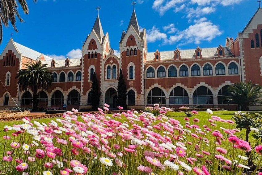Un edificio grande con fachada roja y arcos blancos, rodeado por un campo de flores rosadas y blancas. 1