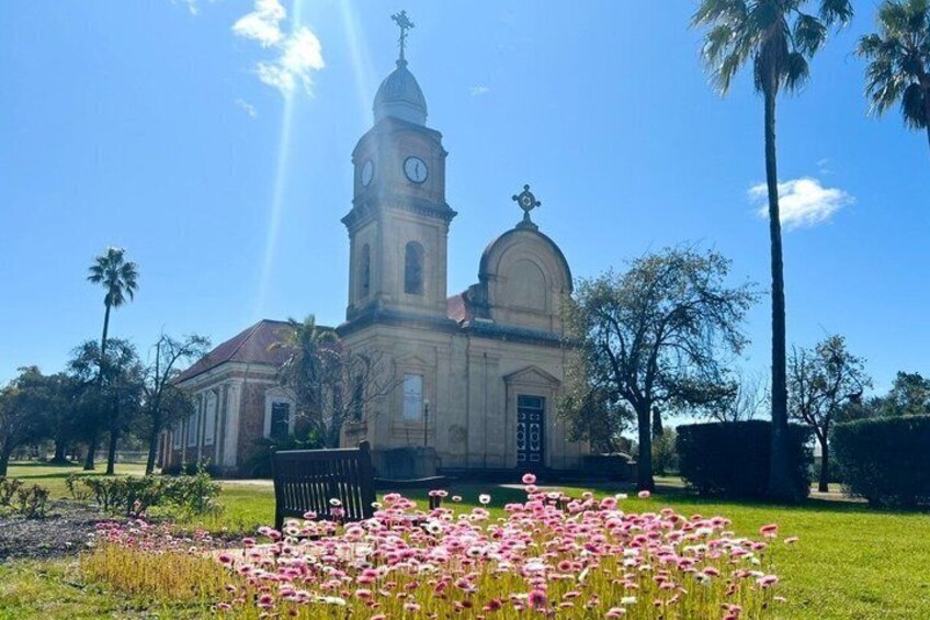 Een kerk met een klokkentoren, omgeven door een tuin met roze bloemen, palmbomen en een helderblauwe lucht. 3