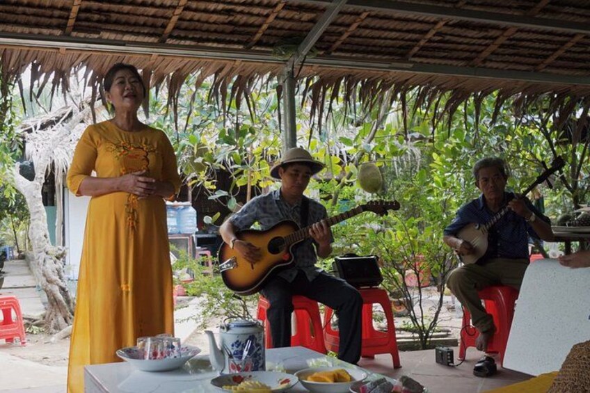 A person in a yellow dress standing, two musicians playing guitars, and a tropical setting with trees and a thatched roof. 5