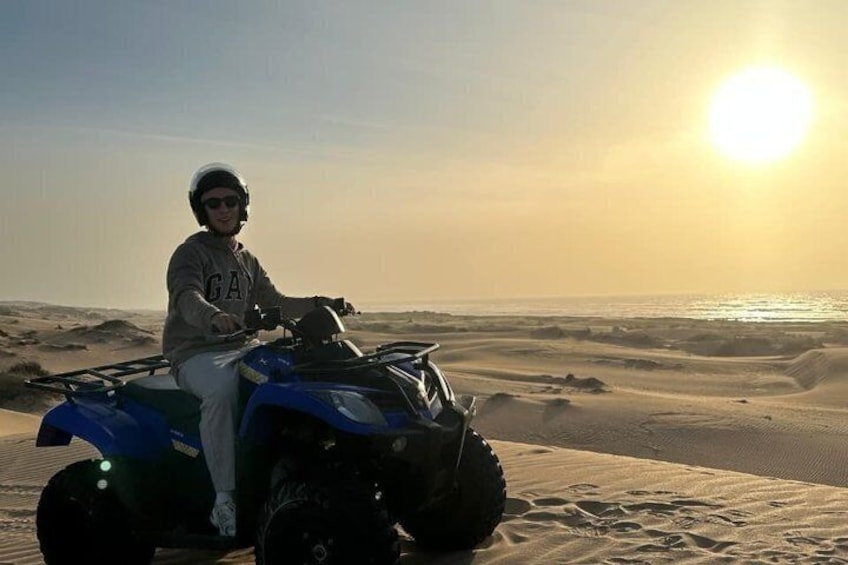 En person på en blå quad bike i et ørkenlandskab med sanddyner og en solnedgang i baggrunden. 2