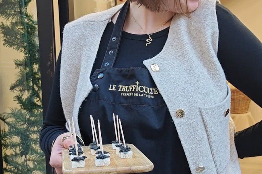 Una persona che tiene un vassoio con piccoli assaggi, indossa un grembiule nero con scritte e un gilet grigio. 3