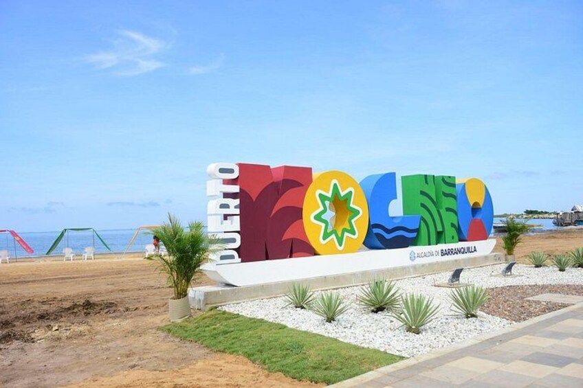 A colorful sign with the text 'PUERTO DE CORUCHO' and a logo, set against a beach backdrop with clear skies. 1