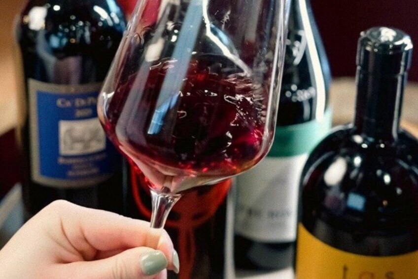 A hand holding a wine glass filled with red wine, with bottles in the background. 3
