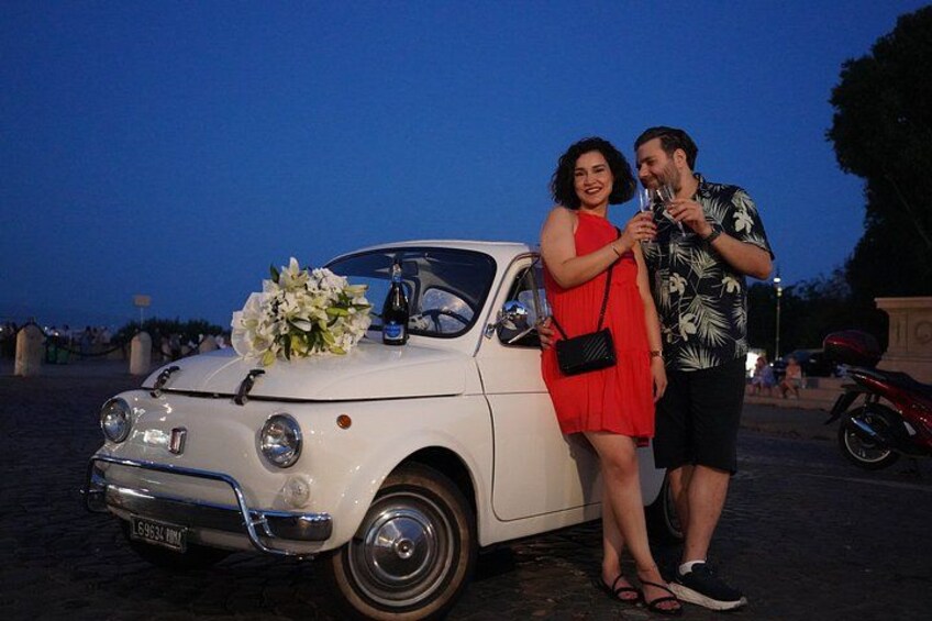 A white vintage car with a bouquet of flowers on the hood, parked on a paved area with people in the background. 1