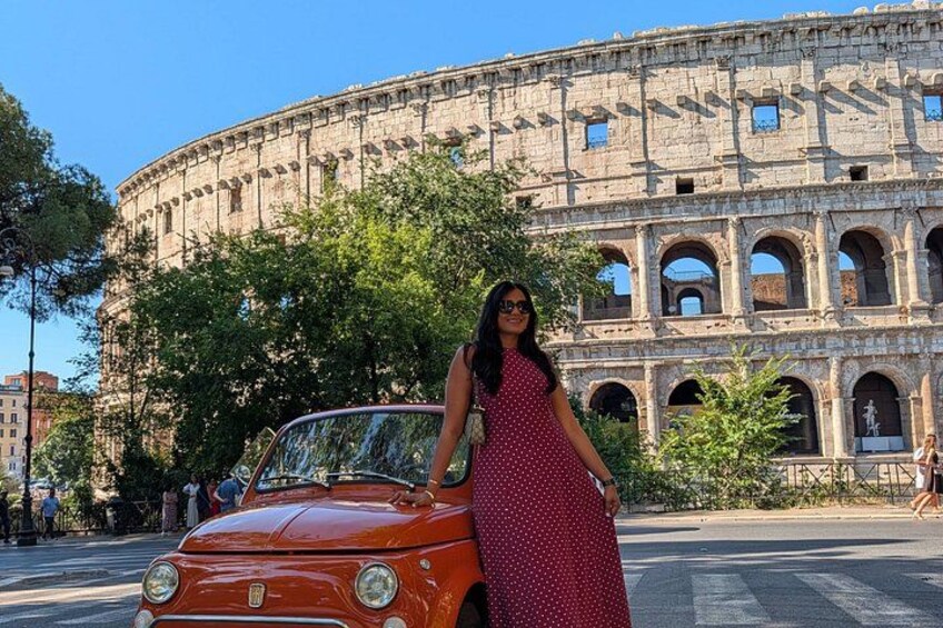 A woman in a polka dot dress standing next to a vintage car with the Colosseum in the background. 3