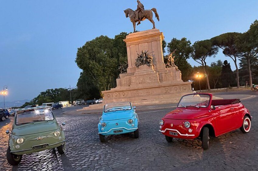 Three vintage cars parked on a cobblestone street with a monument in the background. 4