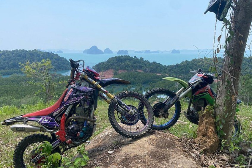 To motocrosscykler parkeret på en jordvej med en smuk udsigt over en skov og et vandareal i baggrunden. 1