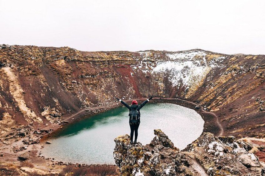 A person standing on a rocky outcrop with arms raised, overlooking a large crater lake surrounded by barren terrain. 5