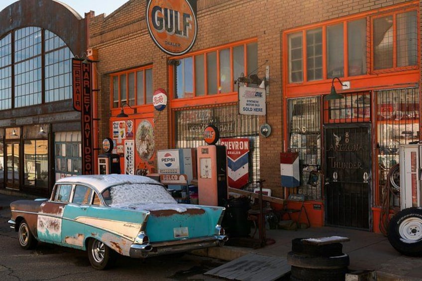 Un coche clásico estacionado frente a un edificio con un letrero que dice 'GULF DEALER', un letrero de 'Harley-Davidson' y un letrero de 'CHEVROLET'. 1
