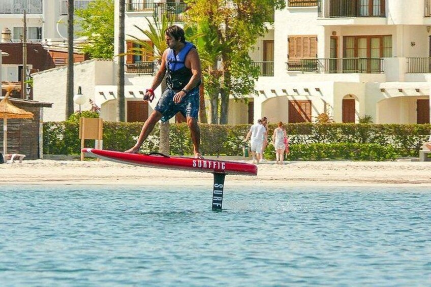 Een persoon surft op een rode plank met de tekst 'SURF' erop, staand op een steiger boven het water, met gebouwen en mensen op de achtergrond. 2