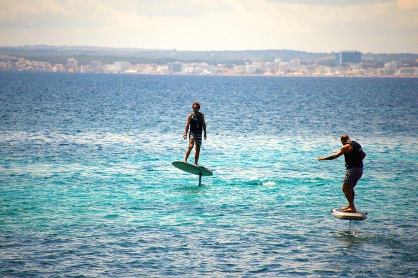 Twee mensen surfen op een kalme zee, met een stadsgezicht op de achtergrond. 1