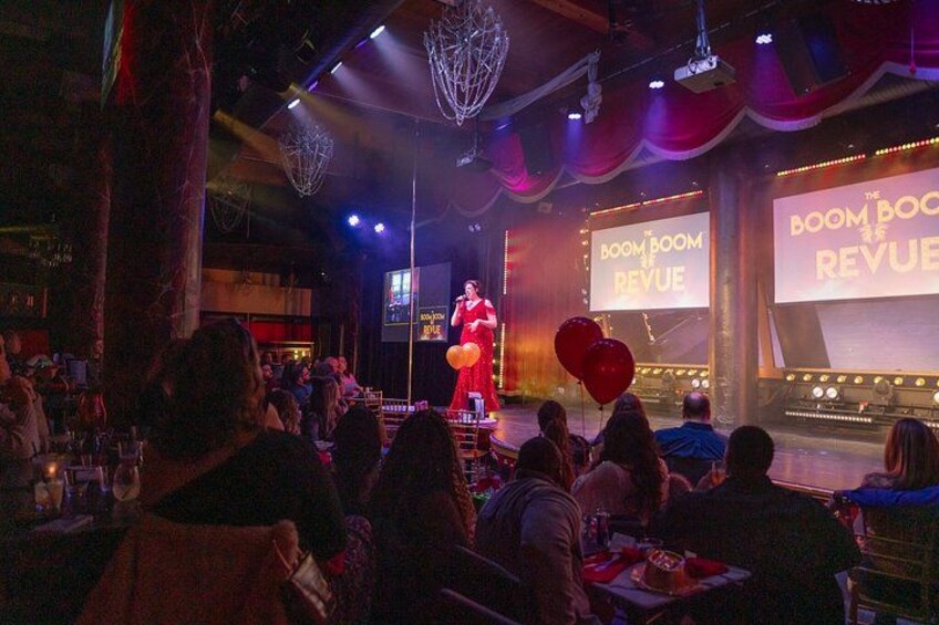 Un escenario con un artista, un público sentado en mesas y una pantalla grande mostrando 'The Boom Boom Revival'. 2