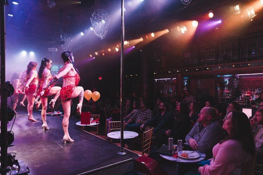 Un escenario con artistas vestidos de rojo, un público sentado en mesas y un bar al fondo. 3