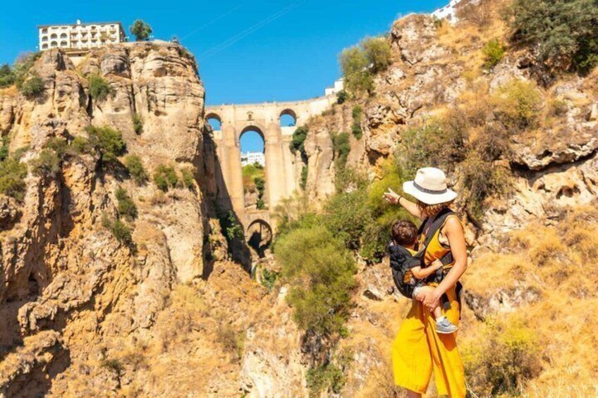 A woman in a yellow dress and white hat holding a baby, standing on a rocky cliff with a historical aqueduct in the background. 2