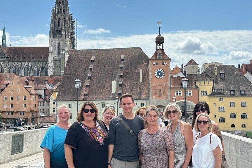 A group of people on a bridge with historic buildings and a clock tower in the background. 1