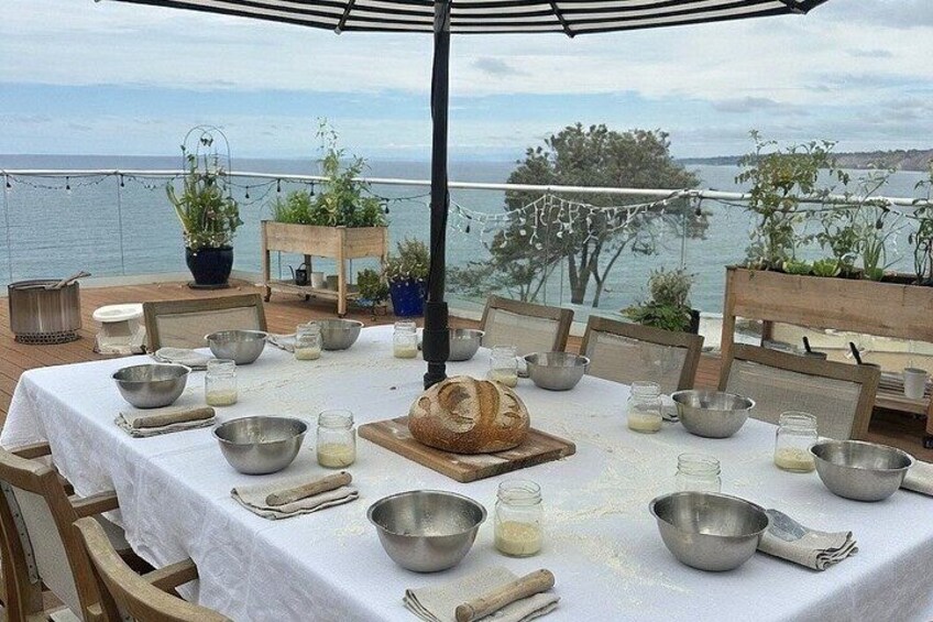 An outdoor dining setup with a table set for a meal, surrounded by chairs, with a view of the sea and plants in the background. 4