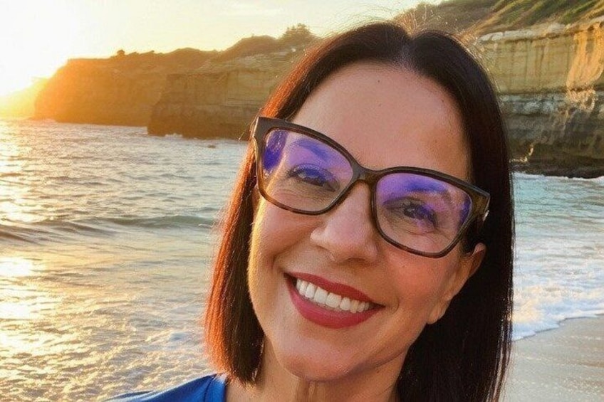 A person with long hair and glasses, standing on a beach with the ocean and cliffs in the background. 2
