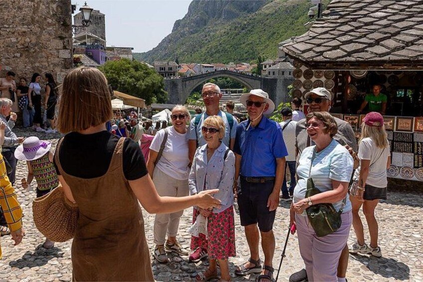 A group of tourists in a historical town square with stone buildings and a bridge in the background. 3