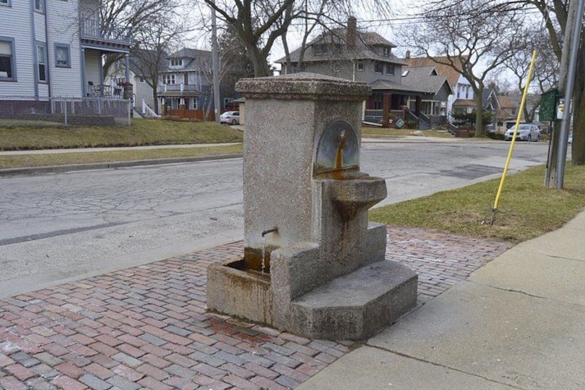En väderbiten stenfountain med en staty, placerad på en trottoar av tegelstenar intill en väg. 3
