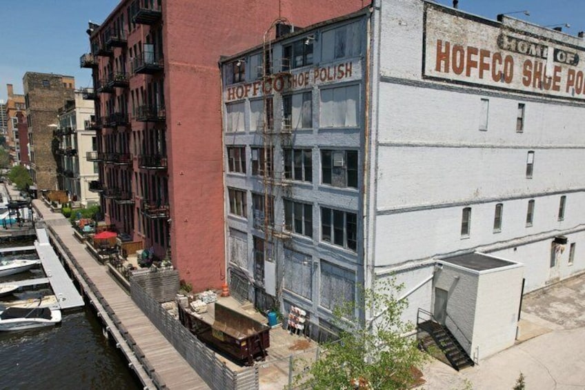 Un bâtiment situé au bord de l’eau, portant une enseigne « HOFFCO SHOE POLISH », un quai avec des bateaux et une rue étroite bordée d’immeubles. 4