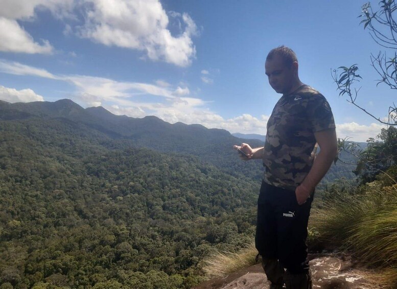 A person standing on a mountain ridge overlooking a dense forest and distant mountains under a partly cloudy sky. 4