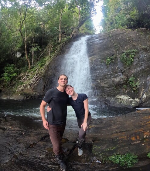 Two people standing on a rock by a waterfall in a forested area. 3