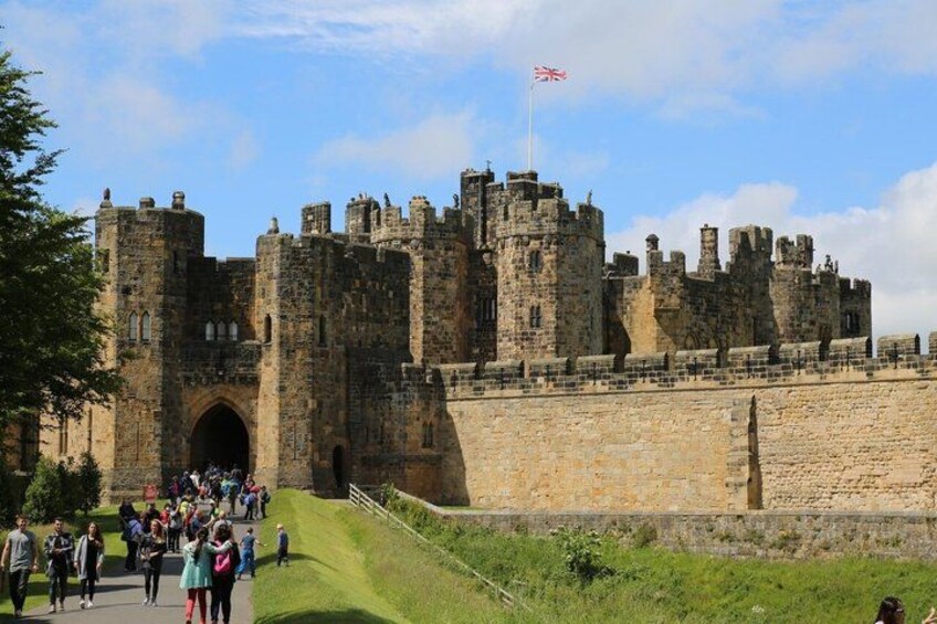 Eine historische Burg mit mehreren Türmen und einer Flagge, umgeben von einer Steinmauer und einer Rasenfläche, auf der Menschen spazieren gehen. 4
