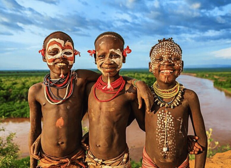 Three individuals in traditional attire with beads and accessories, standing by a river with a clear sky. 1