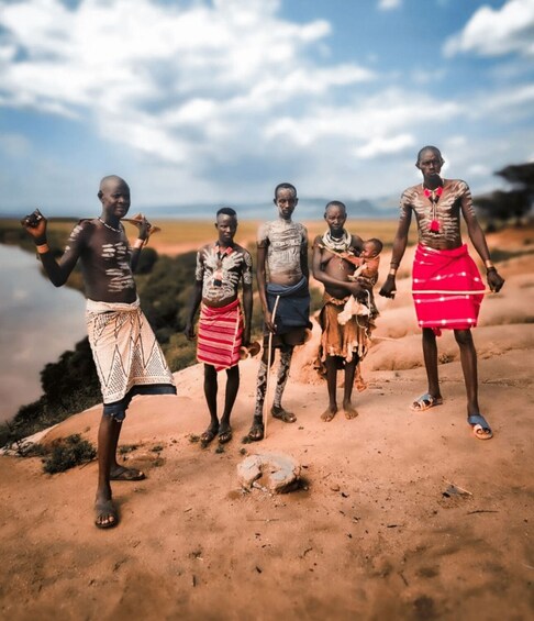 Five individuals in traditional attire standing on a sandy terrain with a body of water and a cloudy sky in the background. 2