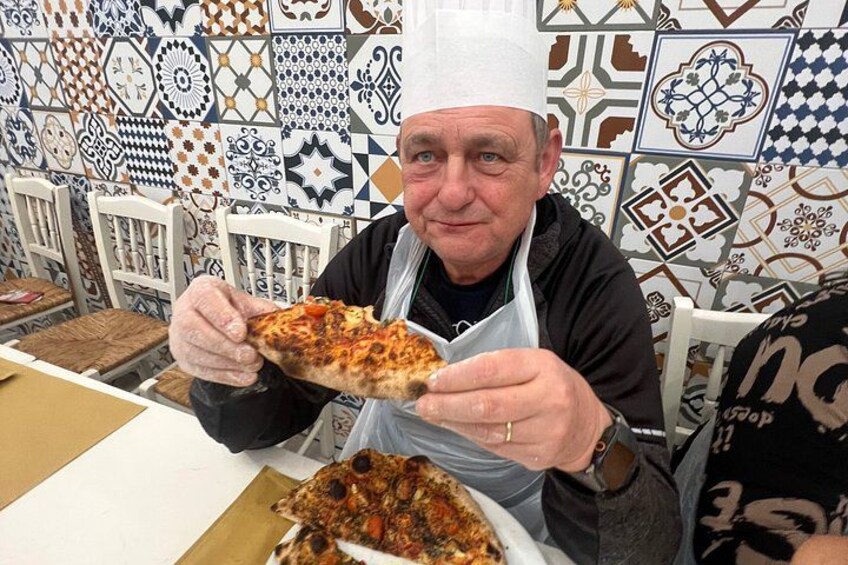 Een persoon houdt een stuk pizza vast, zittend aan een tafel met een wand van betegelde vloertegels op de achtergrond. 2