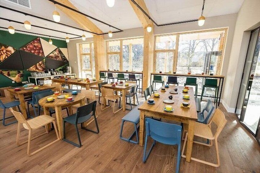 A dining area with wooden tables and chairs, large windows, and a green wall with a food display. 1