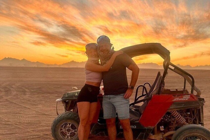 A desert landscape with a sunset, a dune buggy, and two people embracing. 1